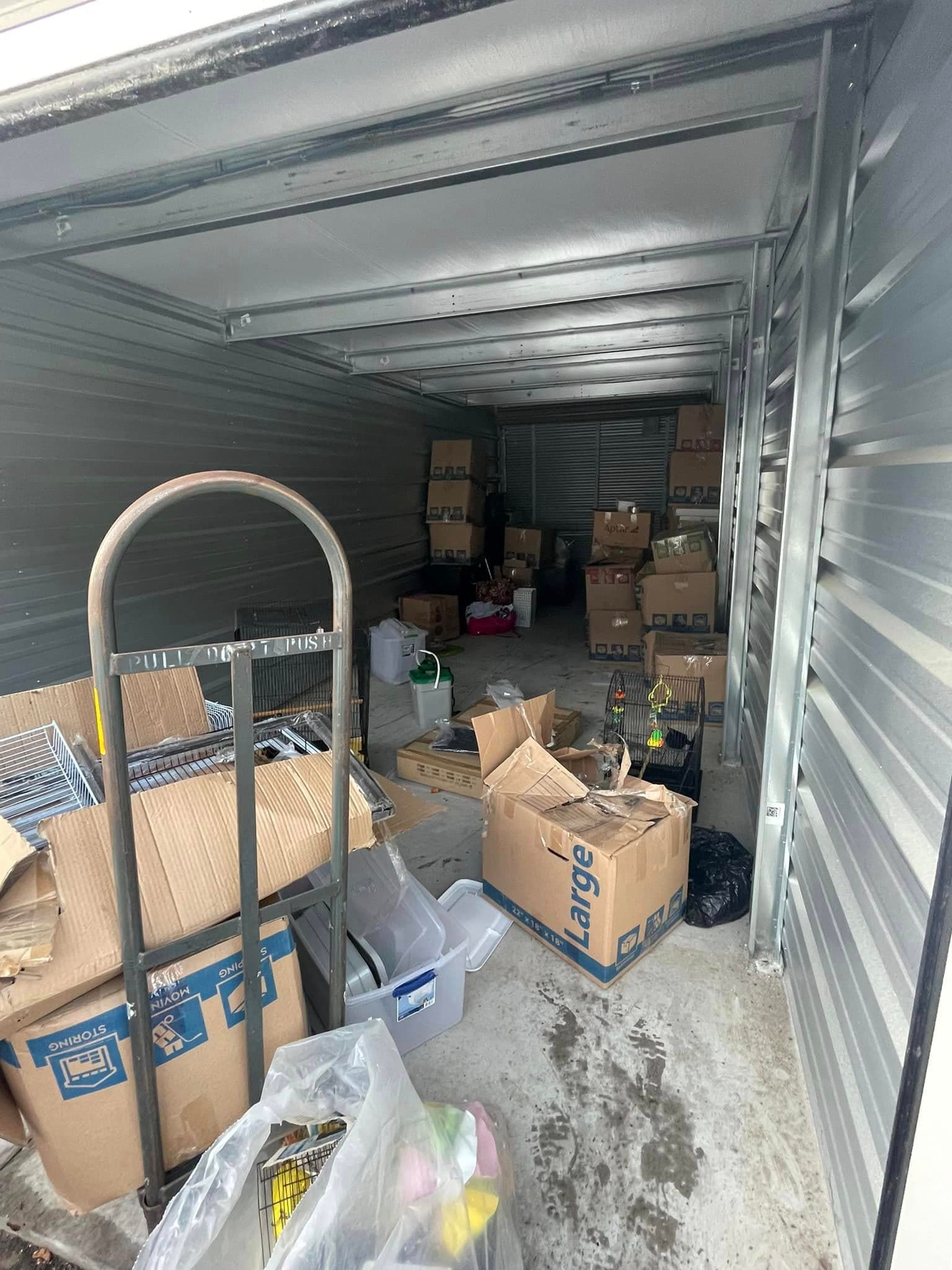 Cluttered storage unit filled with boxes, a cart, and various items scattered around.