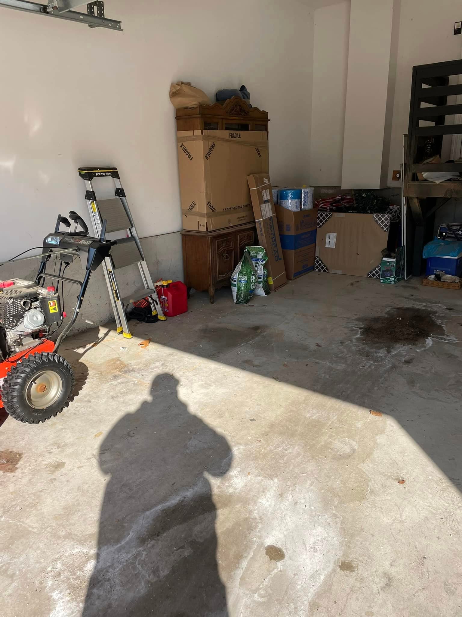 Garage interior with storage boxes, ladder, furniture, and lawn equipment in sunlight.
